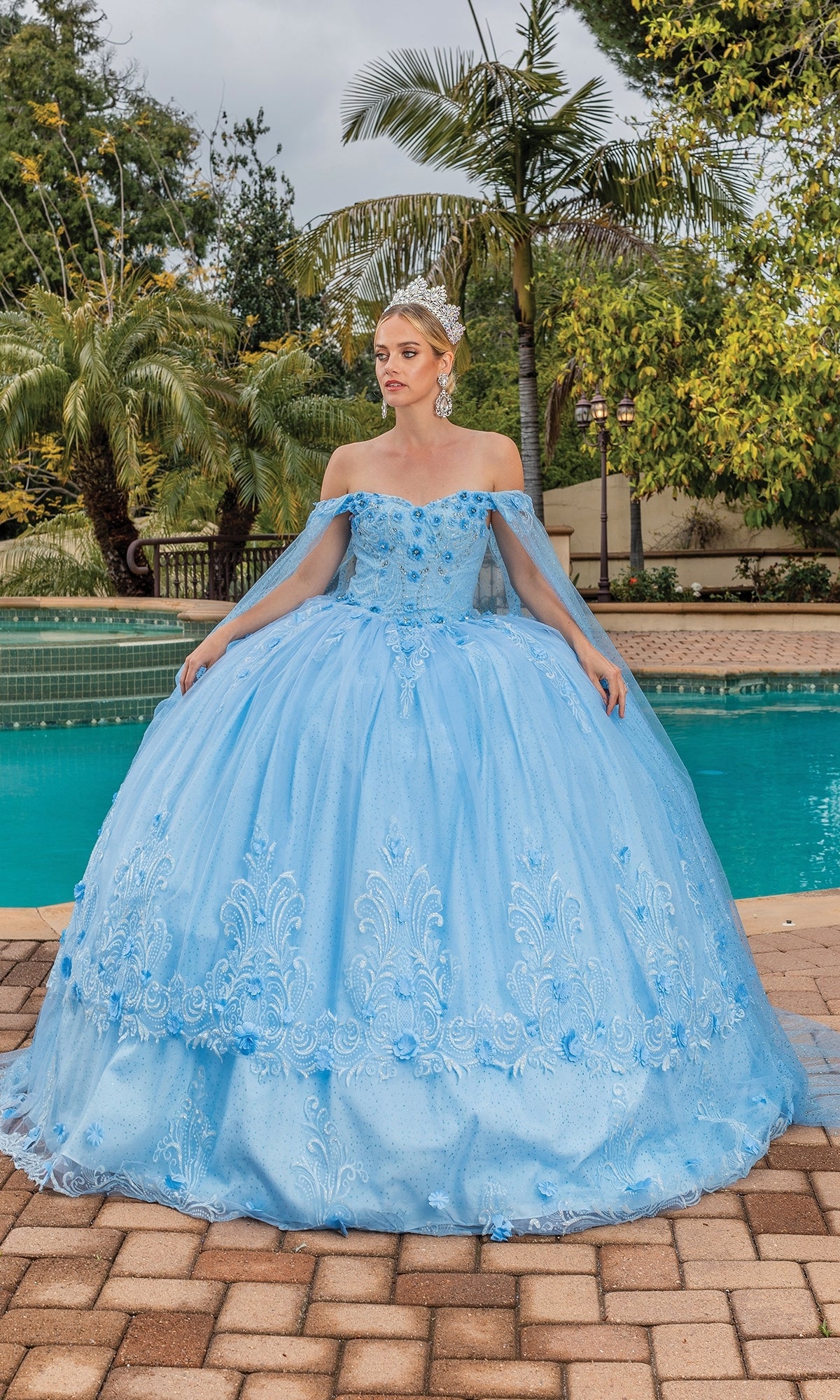 A woman wears the Dancing Queen 1676 Quince Dress with Cape Sleeves in light blue, standing on a paved area by a pool with lush green trees behind her.